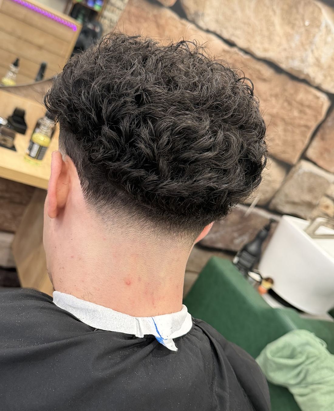 Lockiger Schnitt mit Fade im Nacken, im Hintergrund die Natursteinwand des Salons
